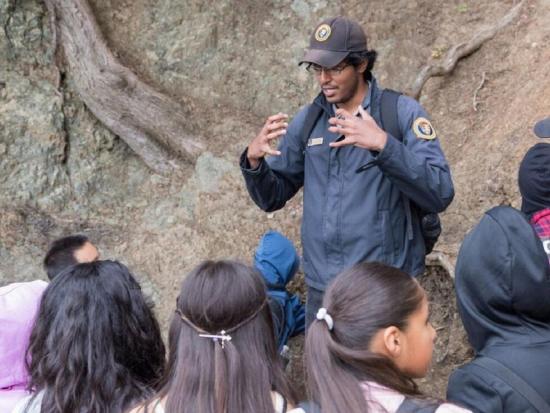 A young man in a volunteer uniform speaks to a group of kids outside.