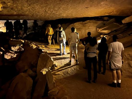 A dozen people stand in a line on a bumpy dirt trail in a dimly lit rock cave.