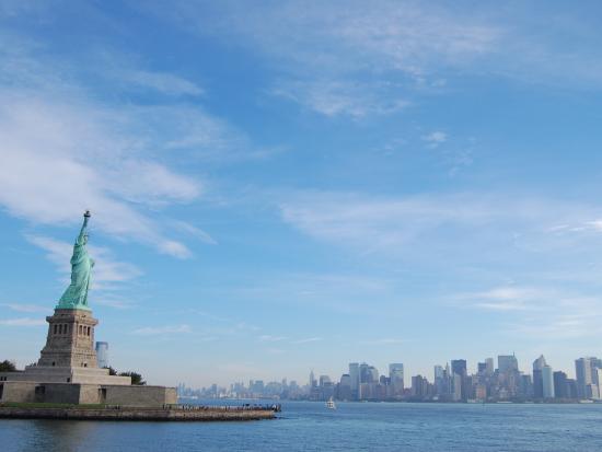 The Statue of Liberty stands on the left side of photo in the middle of body of water with the city skyline in the background