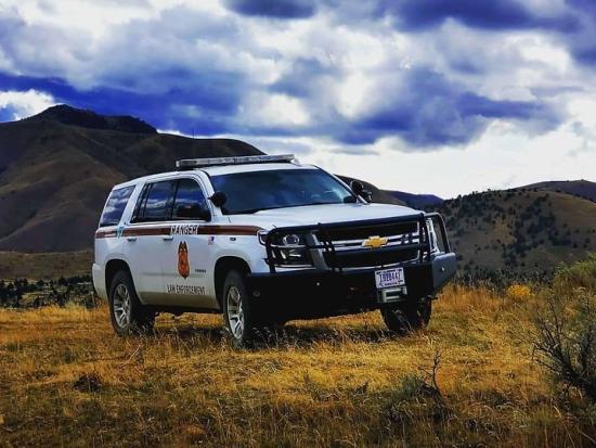 White police car in a field in front of mountains and a cloudy blue sky.