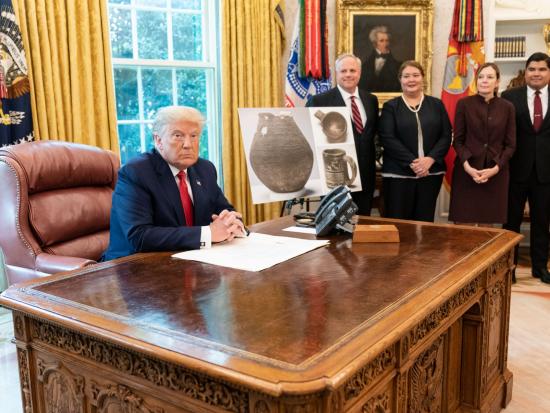 President Trump sits at his desk in the Oval Office with a small group of people and displays of Native American pottery.
