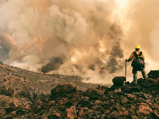 Firefighter stands on a rocky hill, looking toward billowing smoke.