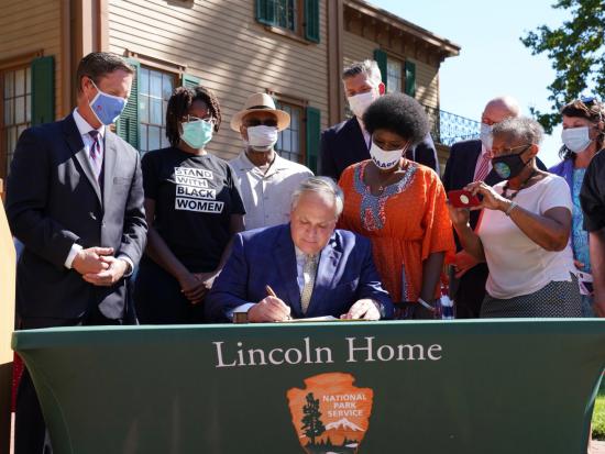 Secretary Bernhardt at a table signing a piece of paper in front of the Lincoln Home and masked onlookers behind him.