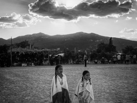 Two Native Americans in a field with mountains behind them and a partly cloudy sky.