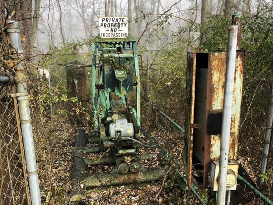 An orphan well with rusted equipment above it surrounded by a fence and brush