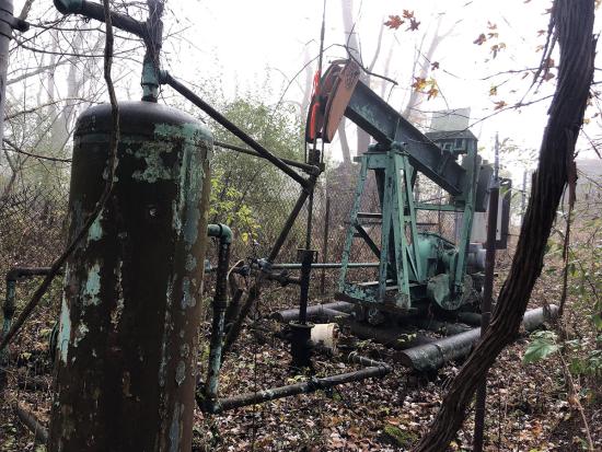 An orphan well with rusted equipment above it surrounded by a fence and brush