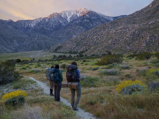Three people wearing backpacks walk along a narrow dirt trail with mountains in the background.