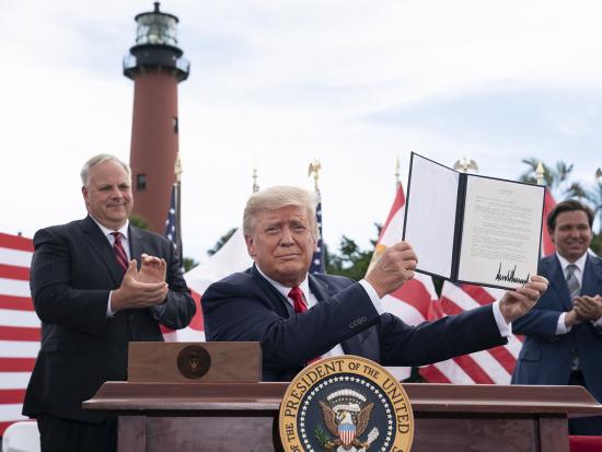 President Trump holds up a signed document while sitting at a table outside with Secretary Bernhardt nearby and a lighthouse in the background.