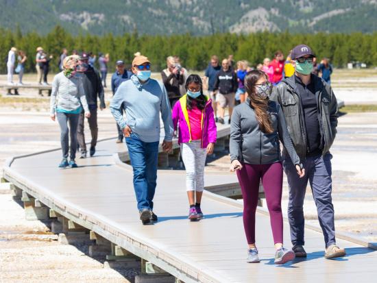 A group of people wearing mask walk along a boardwalk on a sunny day.