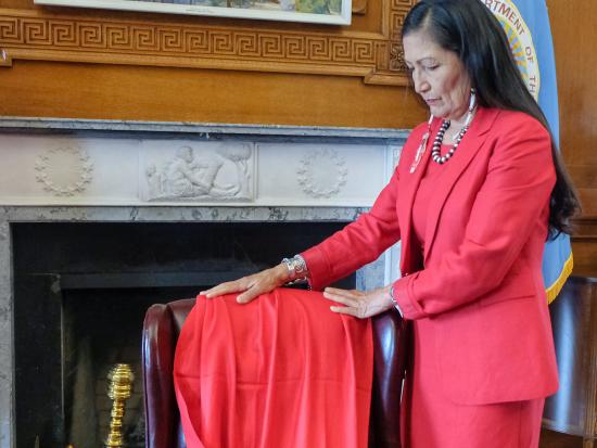 Secretary Haaland solemnly stands next to a red shawl that is on an armchair.