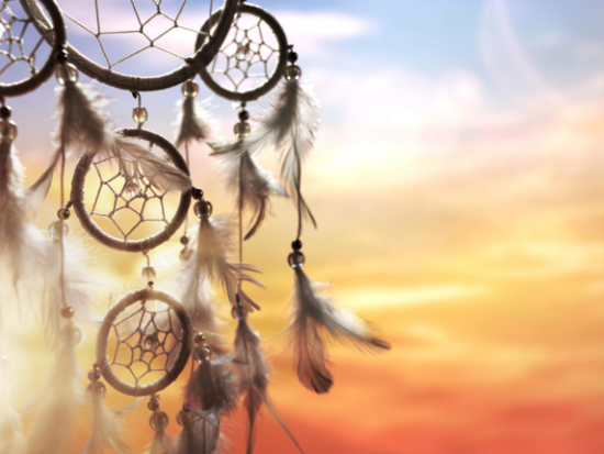 Image of a dreamcatcher with feathers against a sky with the sun setting
