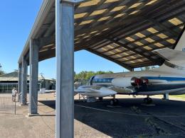 Two people are reading a sign while standing underneath a large open shed housing a jet airplane.