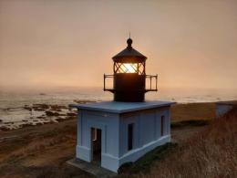 White lighthouse stands lit in front of a sunset over the beach