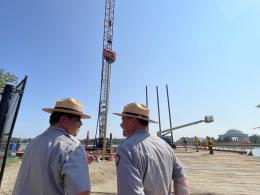In the foreground, two men wearing park ranger uniforms face one another; in the background, a tall piling reaches up and out of frame.