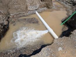 White, frothy water shoots horizontally out of a while plastic pipe into a large muddy puddle in a hole. 