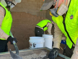 Three people wearing green reflective vests, tan long sleeve shirts, black work gloves, safety glasses and respirators. The two in the front are shoveling coarse sand onto a mesh screen. The third is setting up another screen in the background.