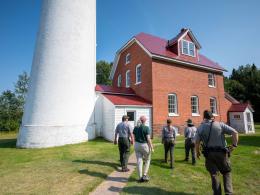 A group of people in National Park Service uniforms stand in front of a lighthouse as they walk along a path on a tour.