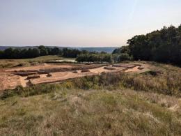 Exposed dirt at building site that is surrounded by green trees in the distance and dry grasses in foreground