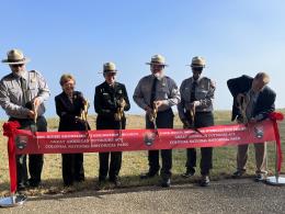 Six people, including four wearing National Park Service uniforms and two in business suits, pose with giant scissors to cut a red ribbon that says York River Shoreline Stabilization Project, Great American Outdoors Act, Colonial National Historical Park