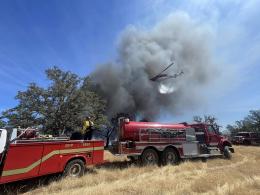 Two red firefighting vehicles are parked in a field of dry grass as smoke rises into a blue sky from a wildfire in the distance. A wildland firefighter stands on one of the vehicles, which is a small wildland fire engine, as he looks towards the rising smoke. A helicopter flies overhead, dropping water from a long hose attached to its belly.