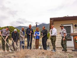 Eight people stand with shovels next to a pile of soil while out in front of a house