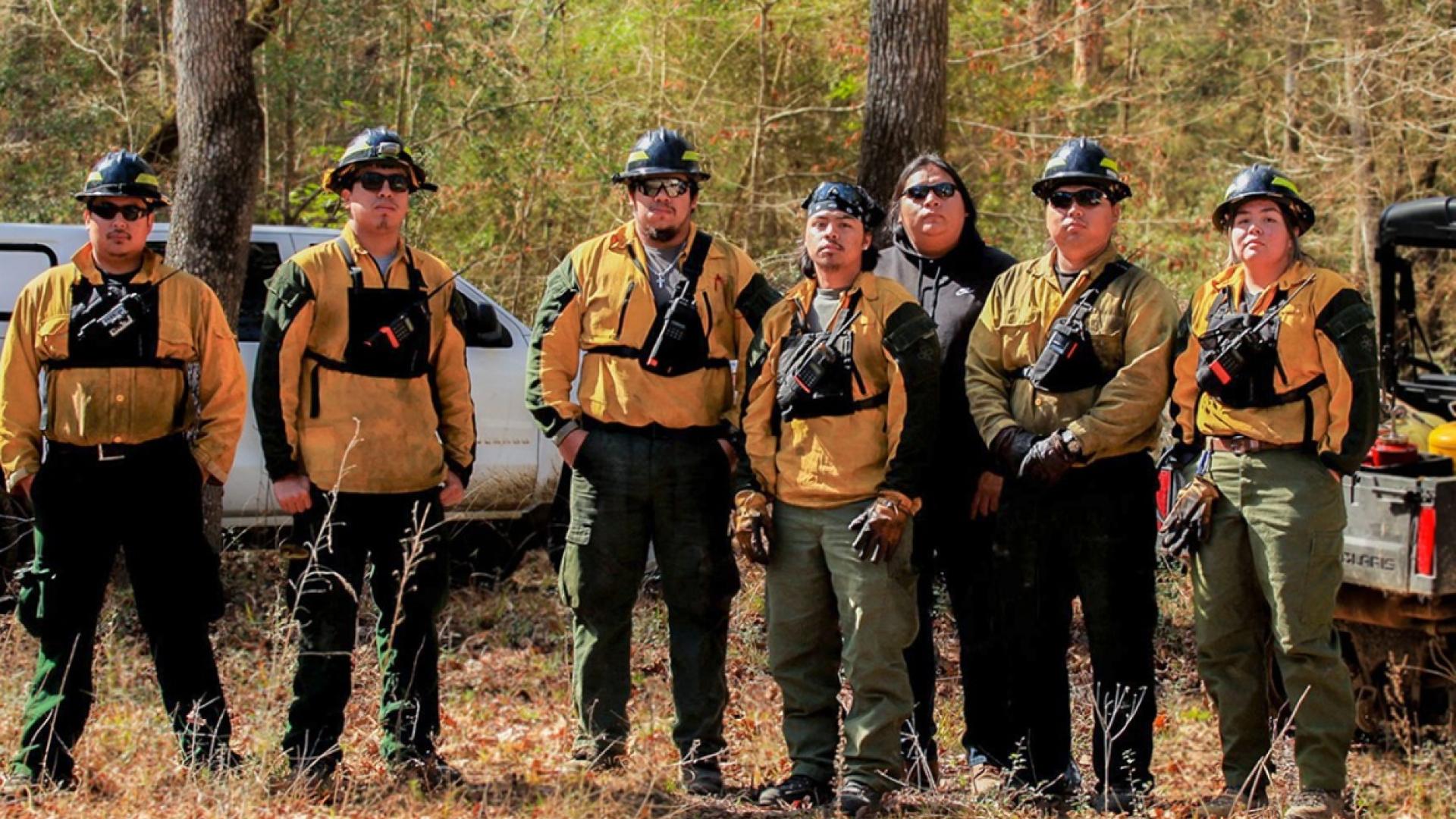 Members of the Alabama-Coushatta Tribe fire management team stand in a leaf-strewn clearing staring at the camera with determined expressions. Behind them is a dense, mixed stand of trees and two vehicles. Photo by Claire Everett, The Nature Conservancy.
