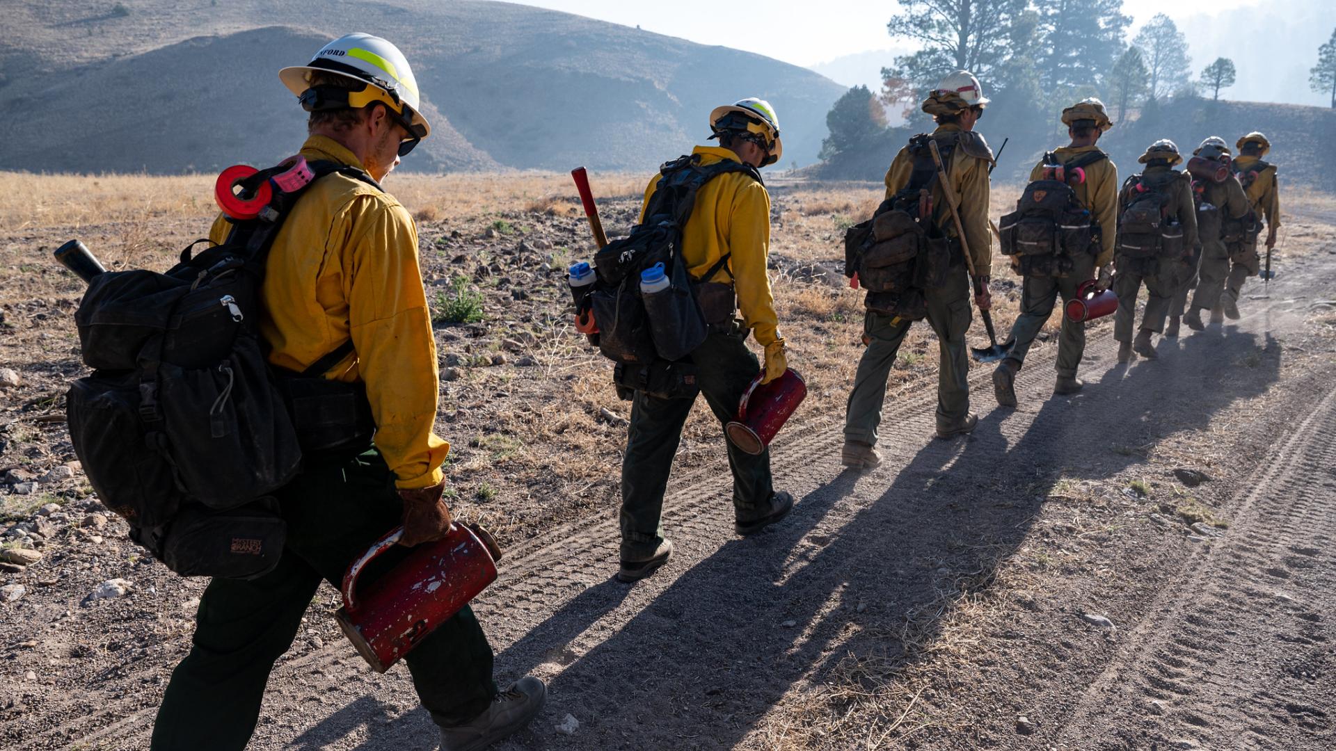 Wildland firefighters hike in a line wearing wildland firefighting gear and carrying tools. 