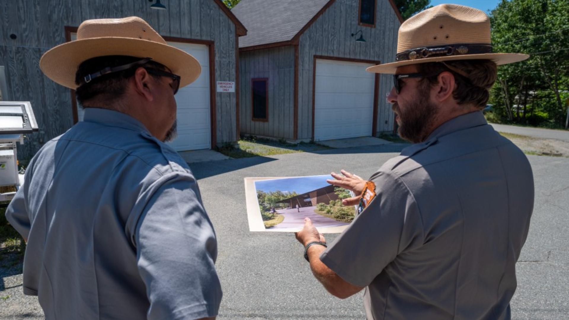 Two uniformed men stand in front of buildings, one holding an image of a building.