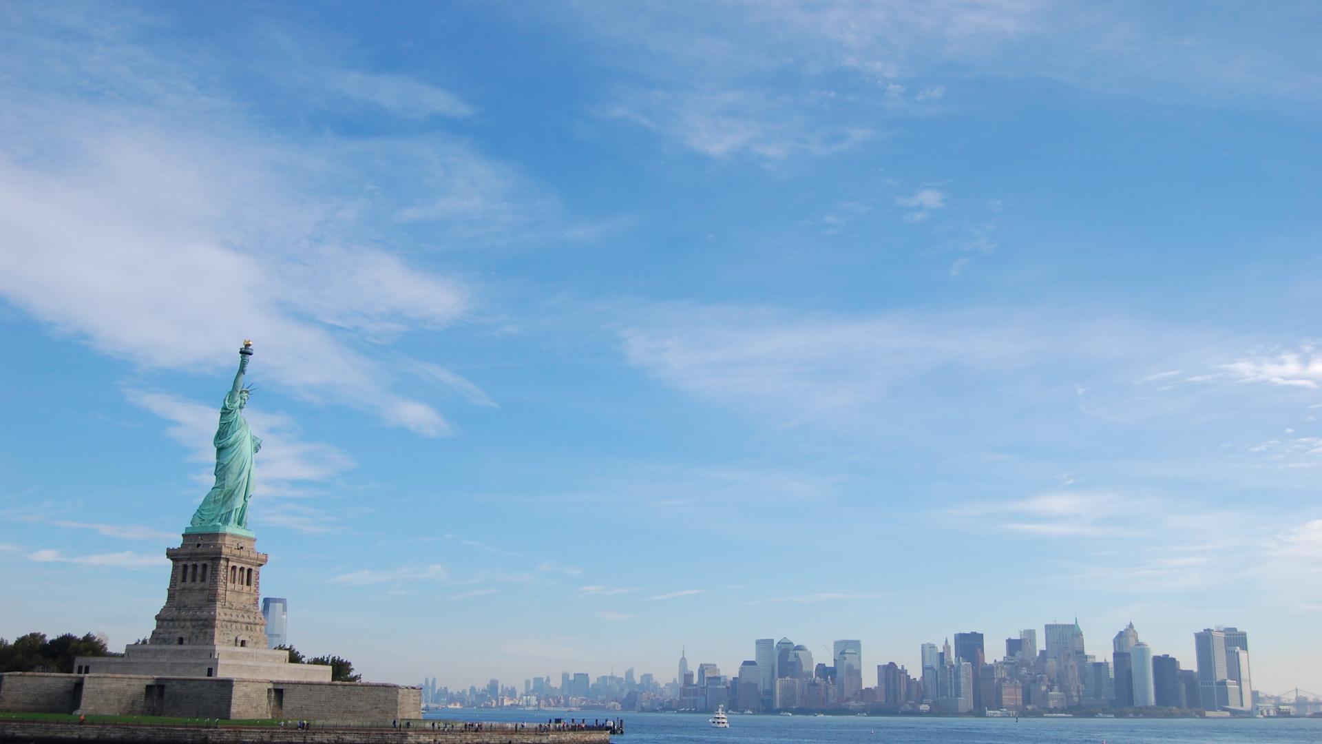 The Statue of Liberty stands on the left side of photo in the middle of body of water with the city skyline in the background