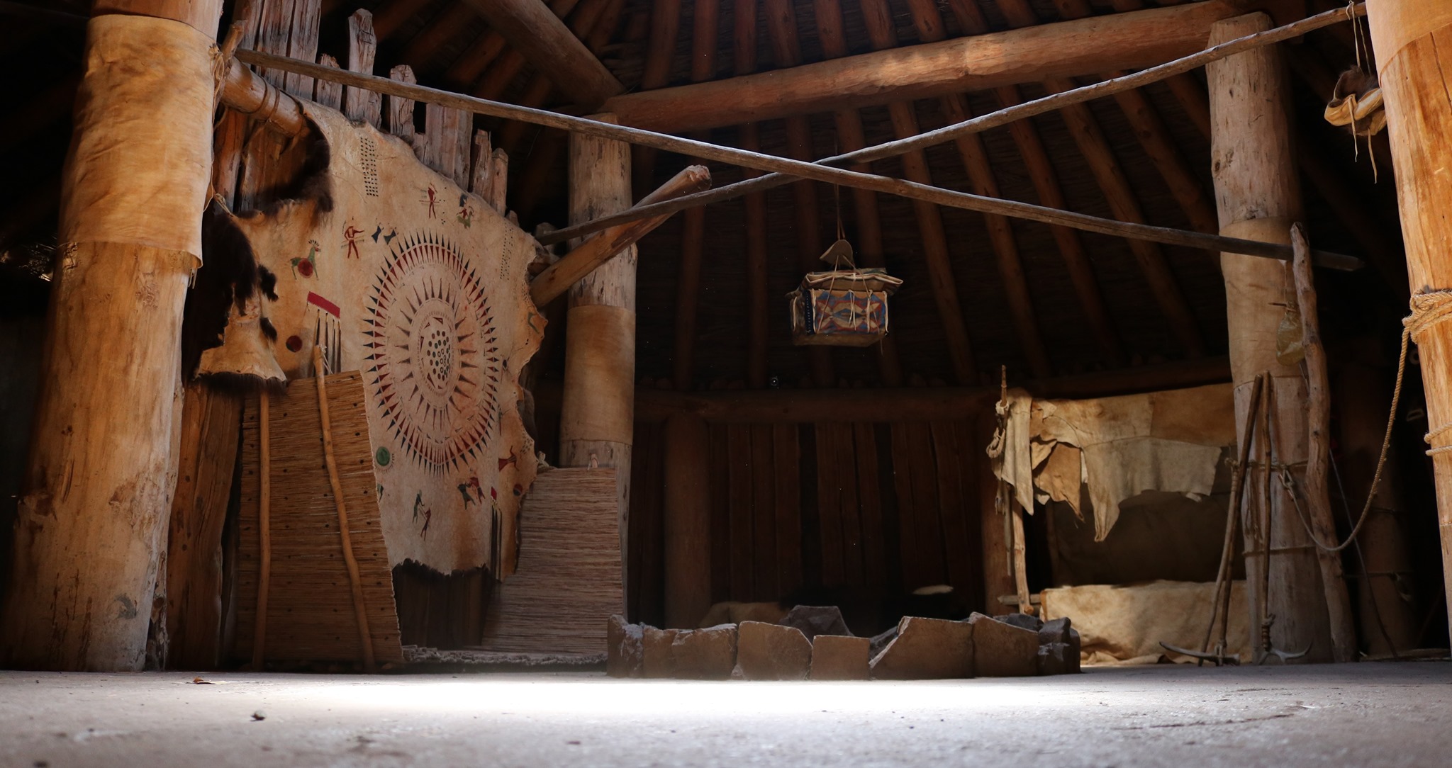 The inside of a wooden lodge is decorated with animal hides and has a fire circle in the middle.
